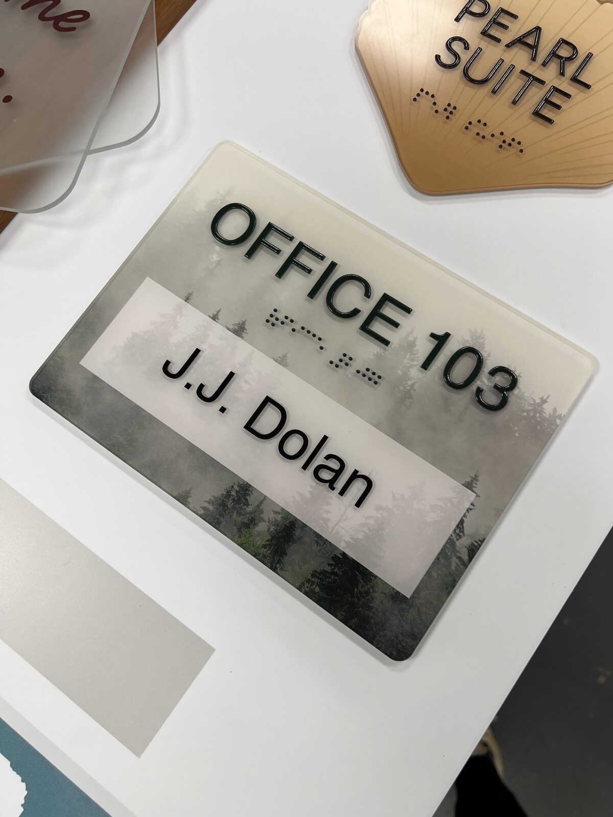 Close-up of an ADA office sign by Studio OntheRock showing raised tactile copy, Grade 2 Braille, and a changeable name insert